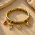 A gold-plated segmented stretch bracelet with three dangling charms: a watermelon slice, a dolphin, and a scallop shell, displayed on a textured beige surface.
