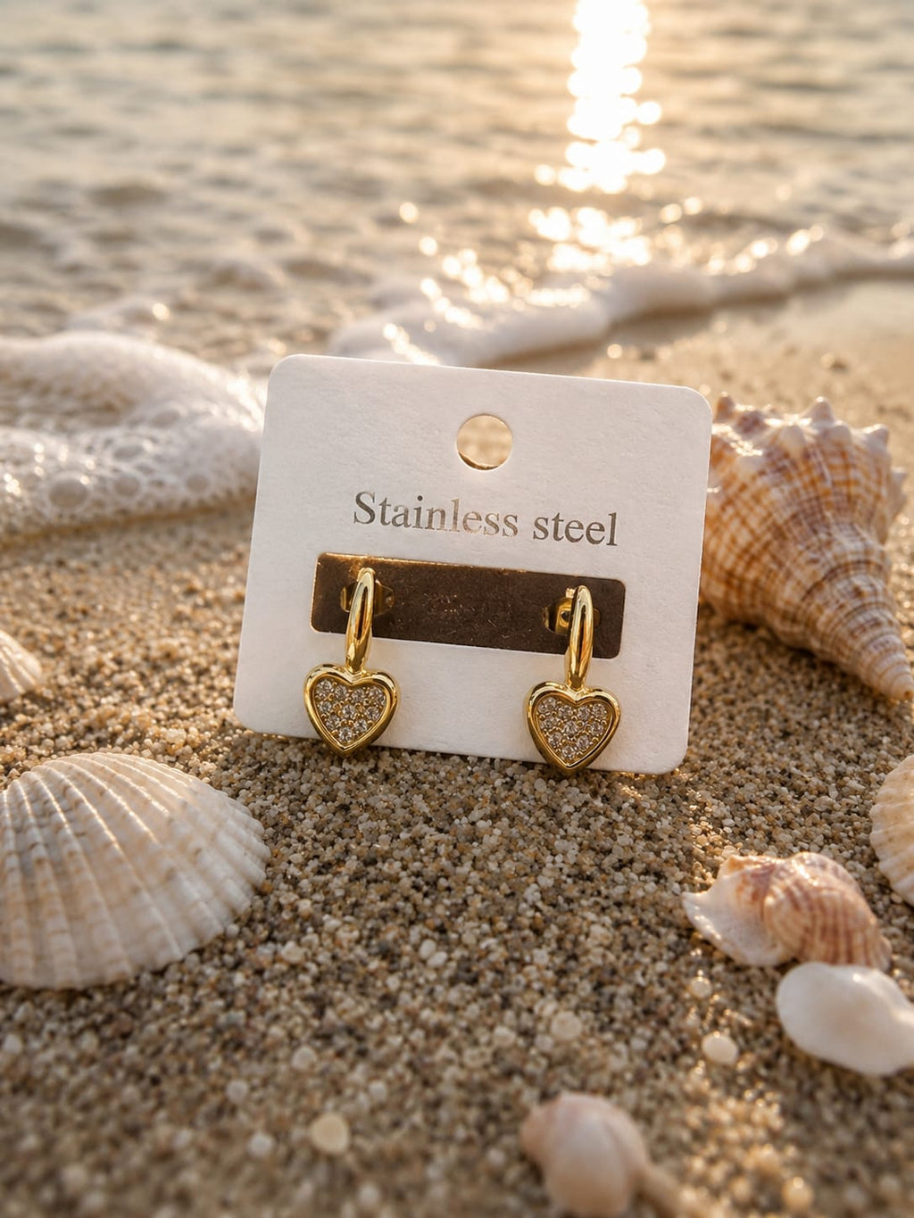 A pair of gold heart-shaped stainless steel dangle earrings displayed on a white card labeled 'Stainless steel', set on a sandy beach with seashells and water in the background.
