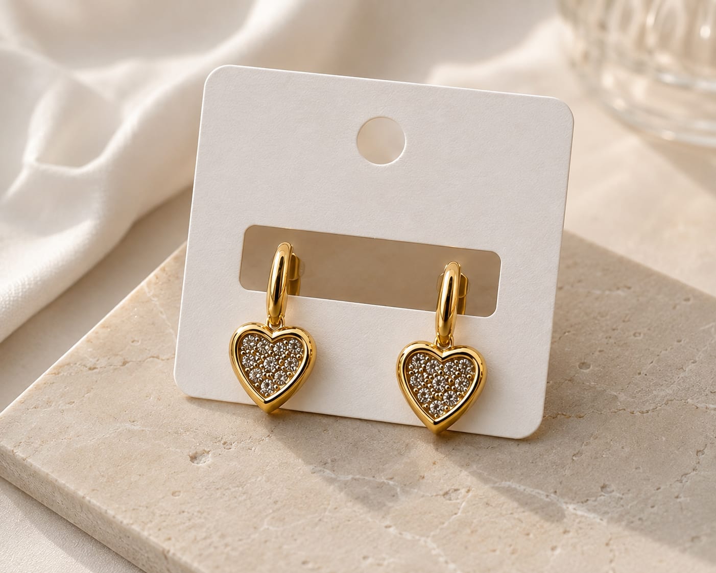 A pair of gold heart-shaped drop earrings with clear pavé-set stones, displayed on a white card hanger over a light stone surface.