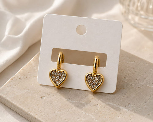 A pair of gold heart-shaped drop earrings with clear pavé-set stones, displayed on a white card hanger over a light stone surface.