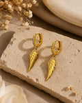 A pair of gold hoop earrings with dangling angel wing charms, decorated with small clear crystals along the hoop, displayed on a textured beige stone block.