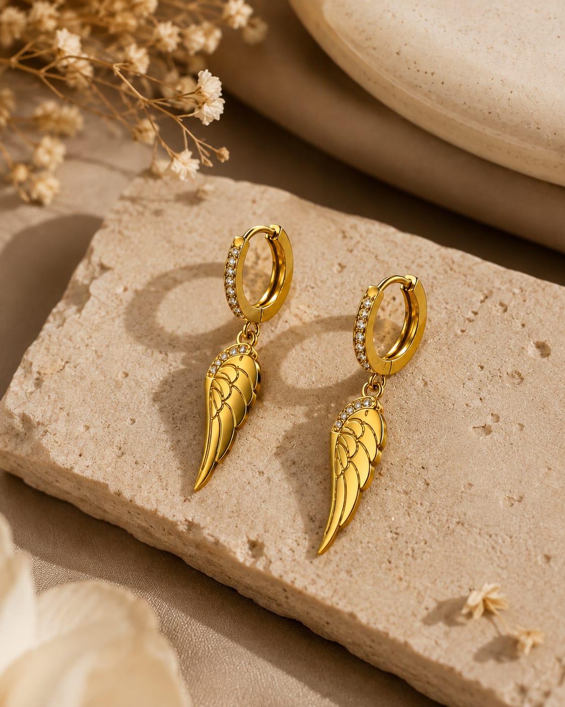 A pair of gold hoop earrings with dangling angel wing charms, decorated with small clear crystals along the hoop, displayed on a textured beige stone block.