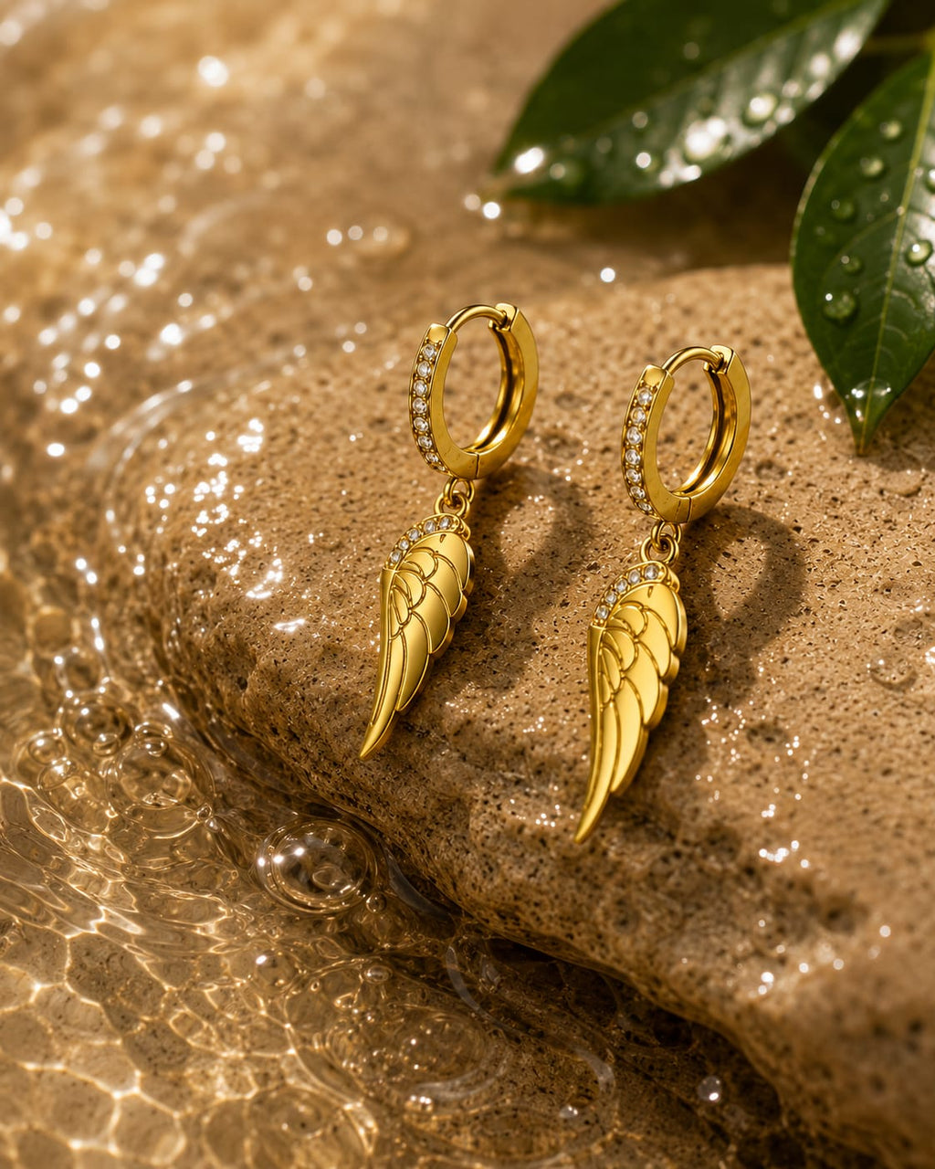 A pair of gold hoop earrings with wing-shaped dangling charms and small clear stone accents, displayed on a wet brown rock with water droplets and green leaves in the background.