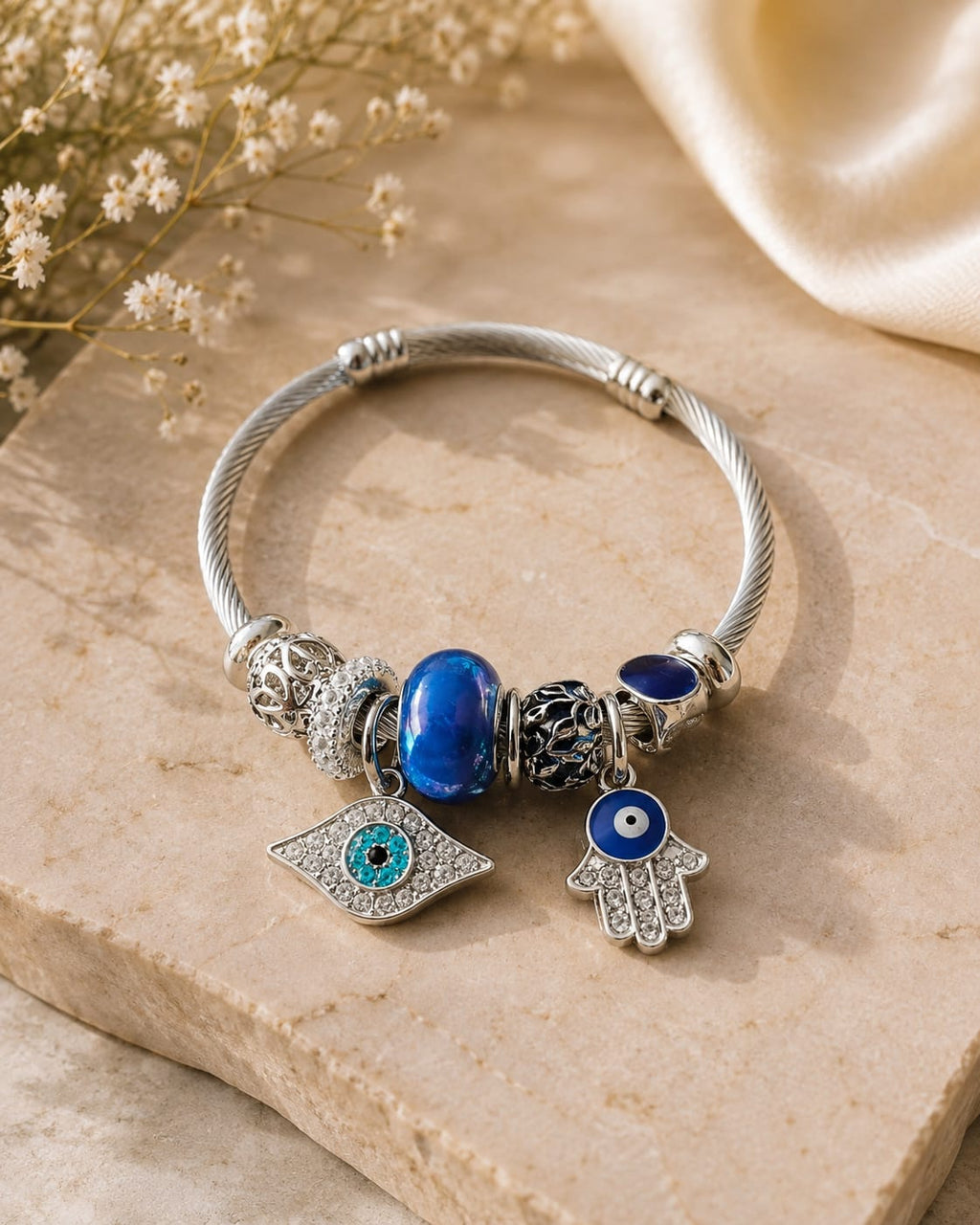 A silver cable-wrapped charm bracelet styled on a beige stone slab with dried flowers and a cream satin cloth. The bracelet features a blue glass bead, a silver evil eye charm, and a silver hamsa hand charm with blue and white details.