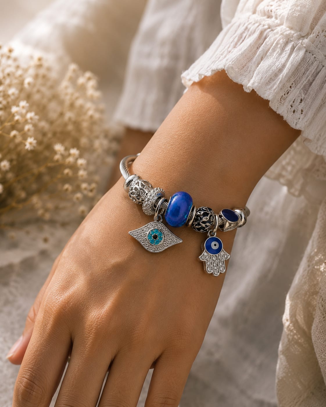 A close-up of a person's wrist wearing a silver charm bracelet with blue and black evil eye beads, a silver hamsa hand charm, and a silver eye of providence charm, styled with a white lace sleeve and dried flowers in the background.
