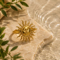 A gold ring shaped like a sun with a human face in the center, displayed on a beige stone slab with water and green leaves in the background.