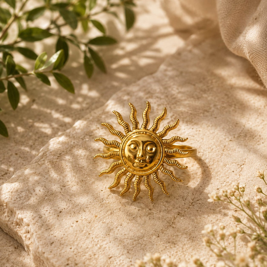 A gold-toned sun face ring with textured rays, displayed on a beige stone surface with soft shadows, green leaves, and a linen corner in the background.