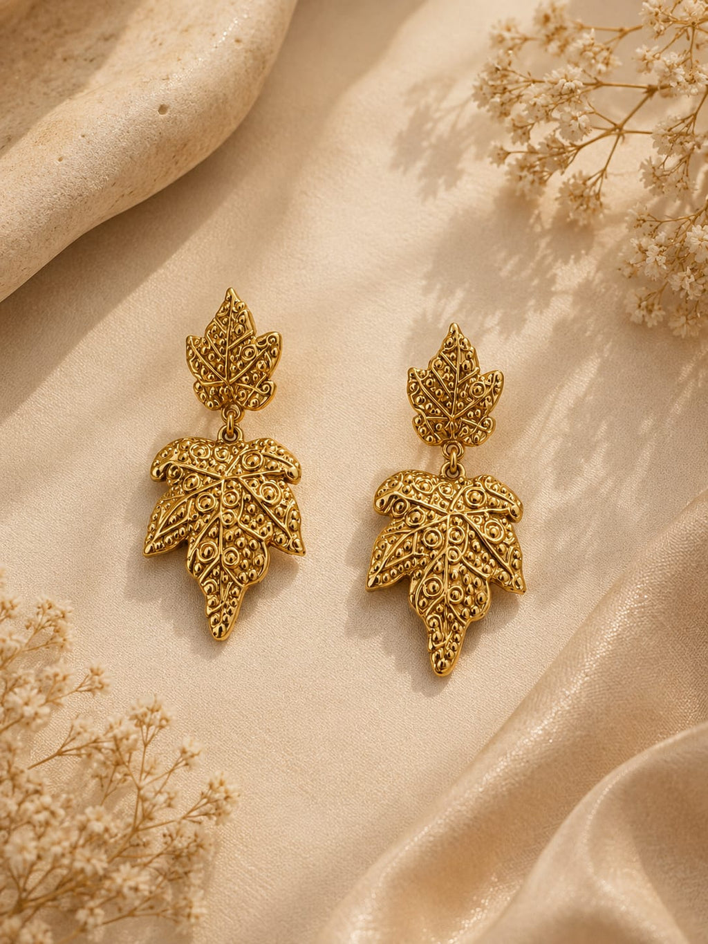 A pair of gold leaf-shaped drop earrings with intricate filigree detailing, displayed on a draped beige fabric background with dried floral accents.