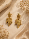 A pair of gold leaf-shaped drop earrings with intricate filigree detailing, displayed on a draped beige fabric background with dried floral accents.