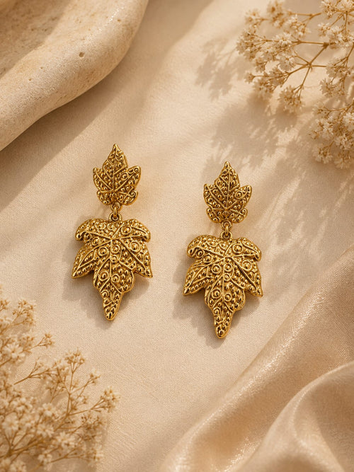 A pair of gold leaf-shaped drop earrings with intricate filigree detailing, displayed on a draped beige fabric background with dried floral accents.