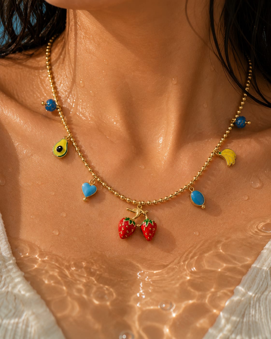 A gold beaded necklace with colorful enamel charms, including two red strawberries at the center, worn on a woman's neck over water.