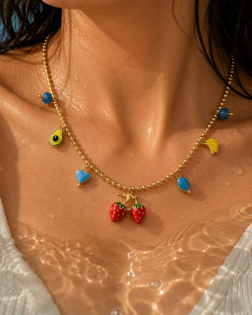A gold beaded necklace with colorful enamel charms, including two red strawberries at the center, worn on a woman's neck over water.