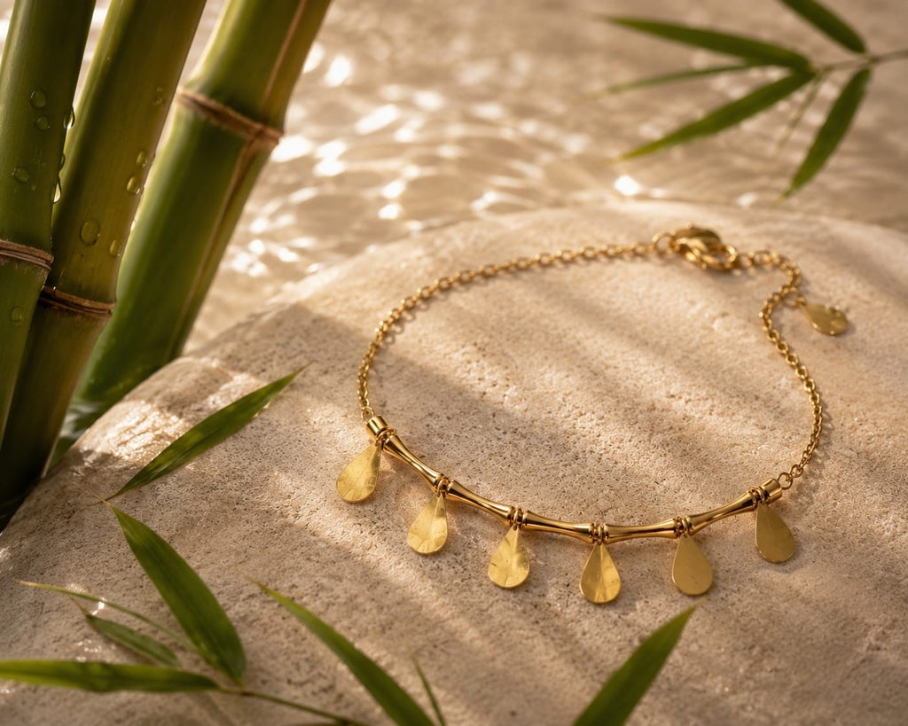 A gold chain bracelet with five teardrop-shaped gold pendants, styled on a textured stone surface with green bamboo leaves in the background.