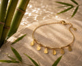 A gold chain bracelet with five teardrop-shaped gold pendants, styled on a textured stone surface with green bamboo leaves in the background.