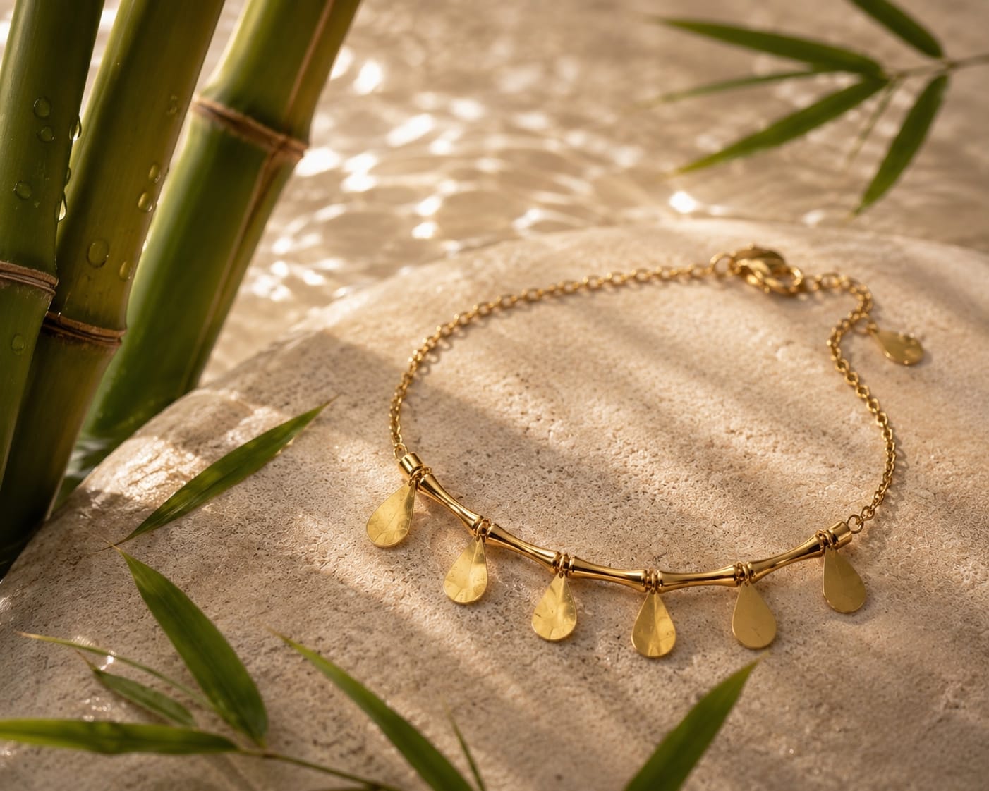 A gold chain bracelet with five teardrop-shaped gold pendants, styled on a textured stone surface with green bamboo leaves in the background.