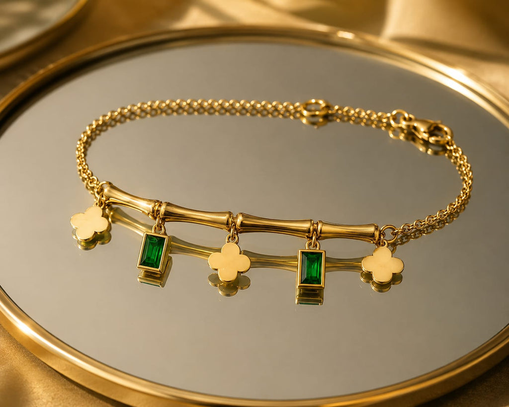 A gold chain bracelet with a bamboo-inspired bar featuring rectangular green emerald-shaped stones and four clover charms, displayed on a reflective gold tray.