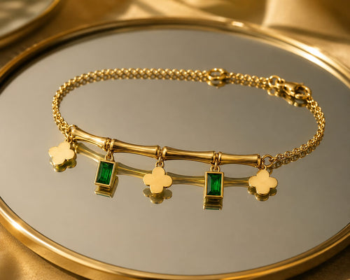 A gold chain bracelet with a bamboo-inspired bar featuring rectangular green emerald-shaped stones and four clover charms, displayed on a reflective gold tray.