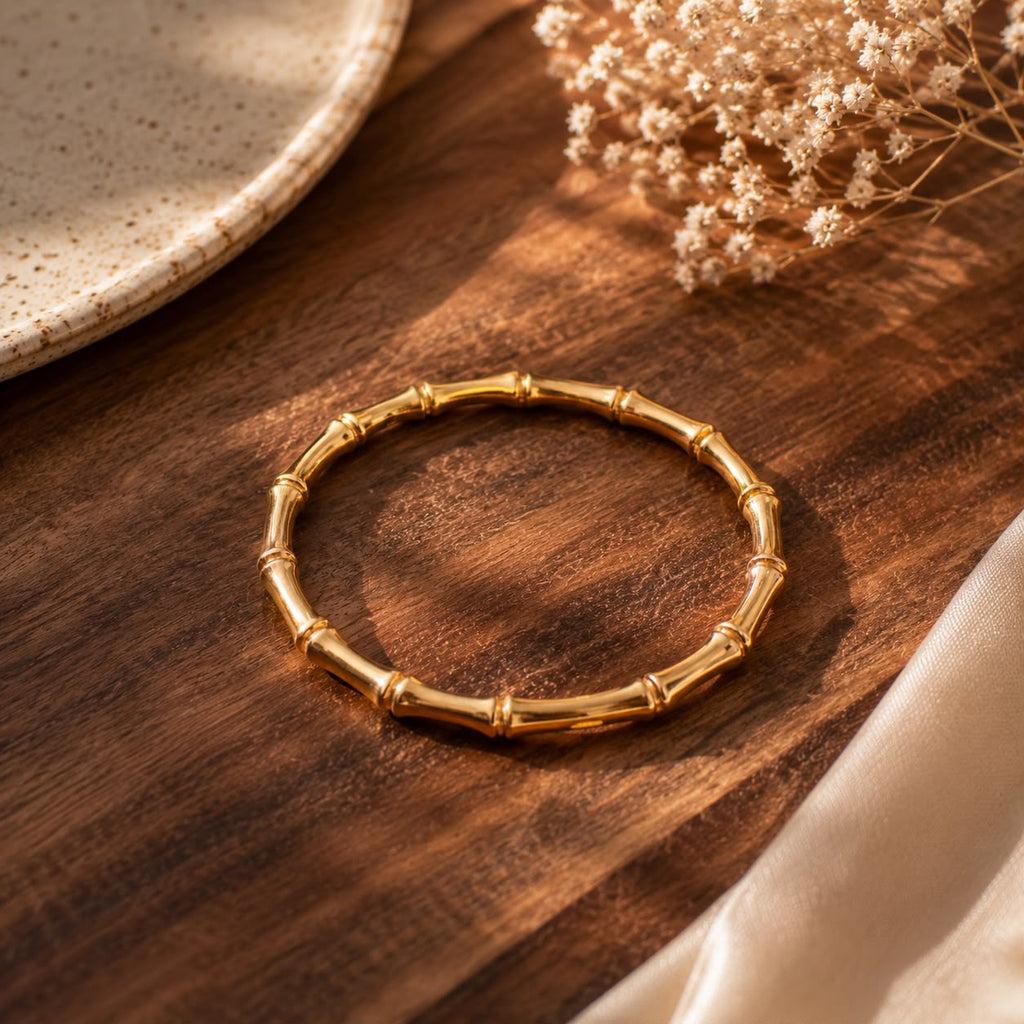 A gold-toned bamboo-style bangle bracelet resting on a dark wooden surface, with a ceramic plate edge and dried flowers in the background.