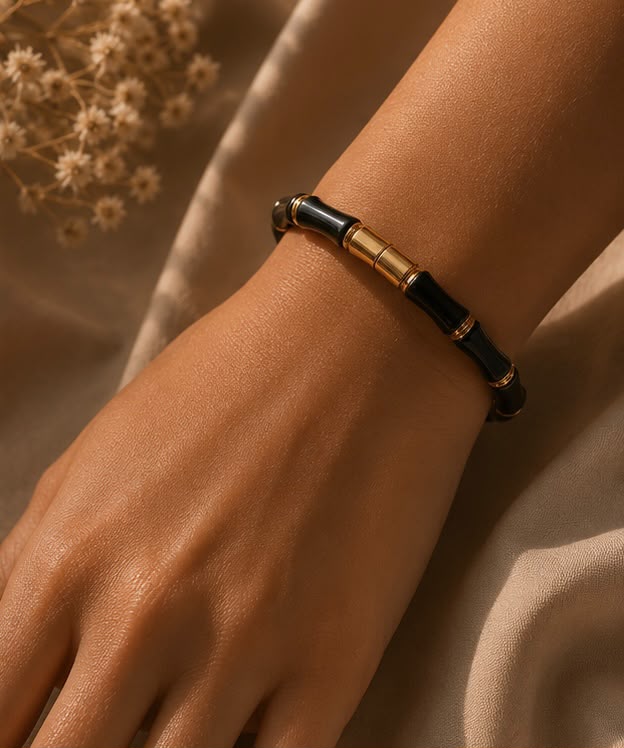 A close-up of a person's wrist wearing a beaded bracelet with alternating glossy black and metallic gold cylindrical beads, set against a soft beige fabric background with dried flowers in the corner.