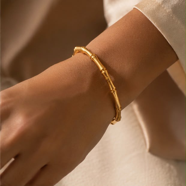 A close-up of a person's wrist wearing a polished gold bangle bracelet with a bamboo joint design, set against a soft beige background.