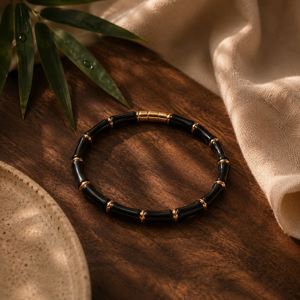 A beaded bracelet with glossy black cylindrical beads and small gold spacer rings, arranged in a circle on a dark wooden surface with beige fabric and green leaves as props.