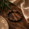 A beaded bracelet with glossy black cylindrical beads and small gold spacer rings, arranged in a circle on a dark wooden surface with beige fabric and green leaves as props.
