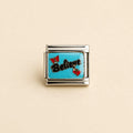 A rectangular silver charm link featuring a bright turquoise blue enamel background with the word 'Believe' in black script, accented by two small red bow charms, shown on a light beige background.