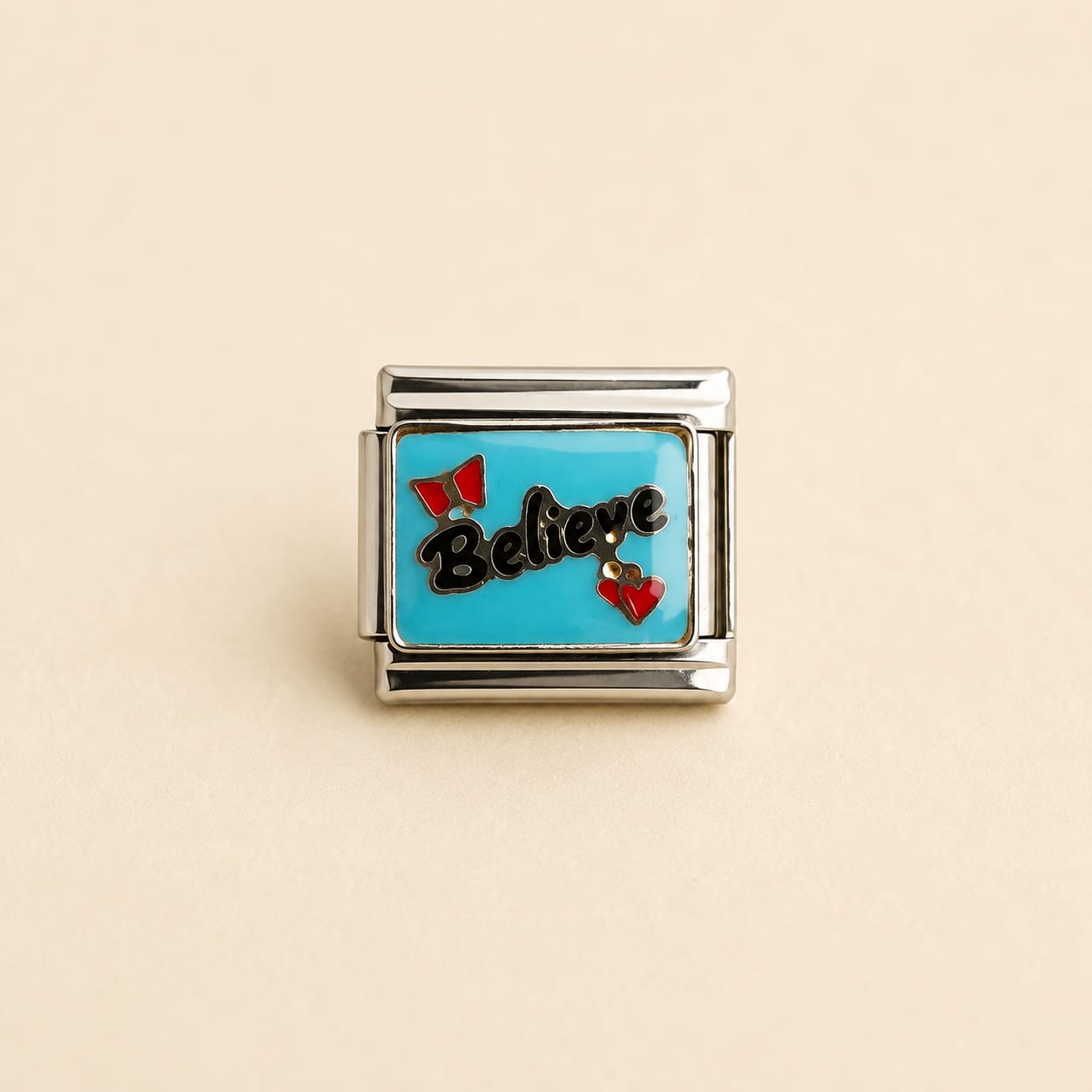 A rectangular silver charm link featuring a bright turquoise blue enamel background with the word 'Believe' in black script, accented by two small red bow charms, shown on a light beige background.