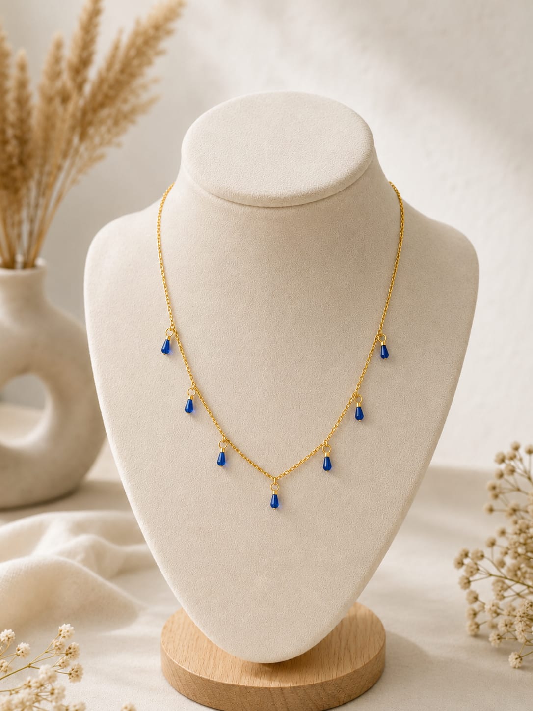 A delicate gold chain necklace with five small blue teardrop charms, displayed on a cream-colored jewelry bust with dried flowers in the background.