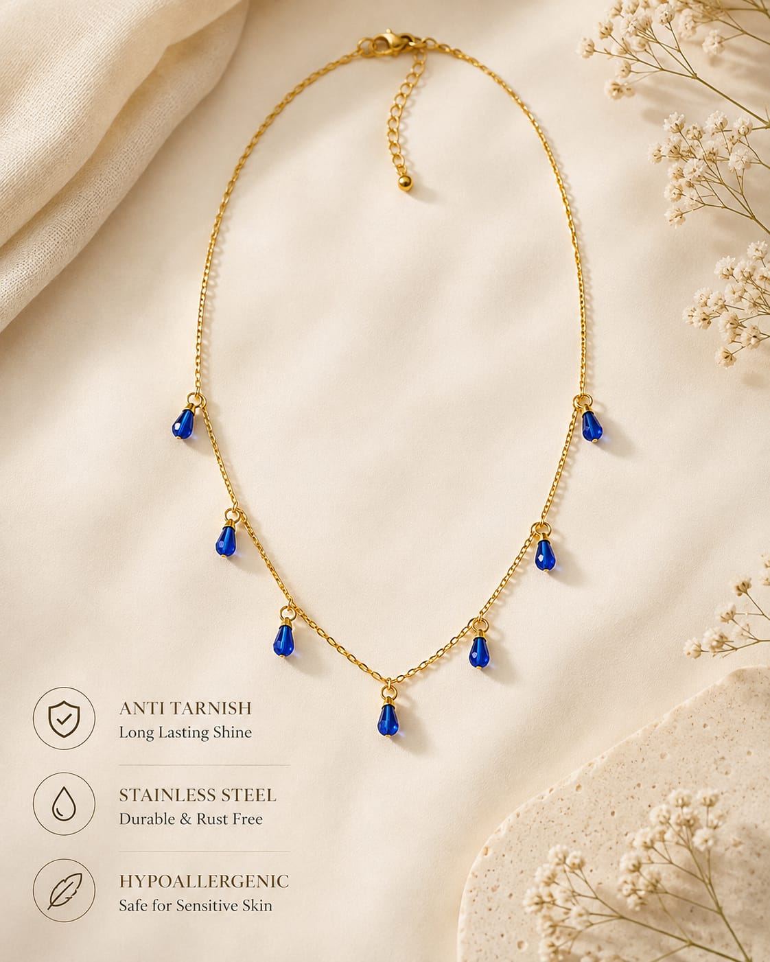 Gold chain necklace with five small blue teardrop-shaped pendant drops, displayed on a soft cream fabric background with decorative dried flowers and three feature callouts on the left.
