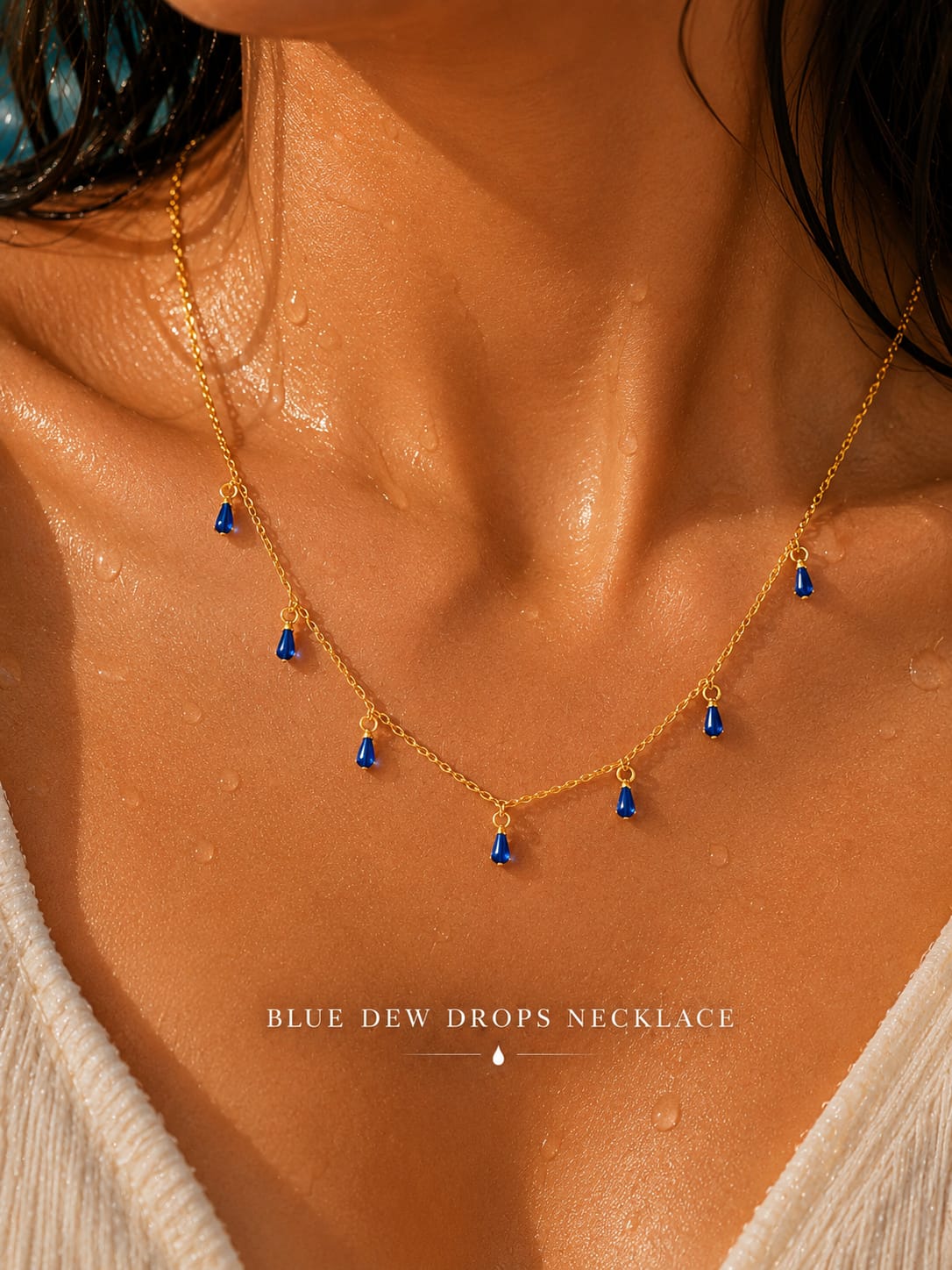 A close-up of a model's neck and chest wearing a delicate gold necklace with five small blue teardrop-shaped drops, labeled 'BLUE DEW DROPS NECKLACE'.