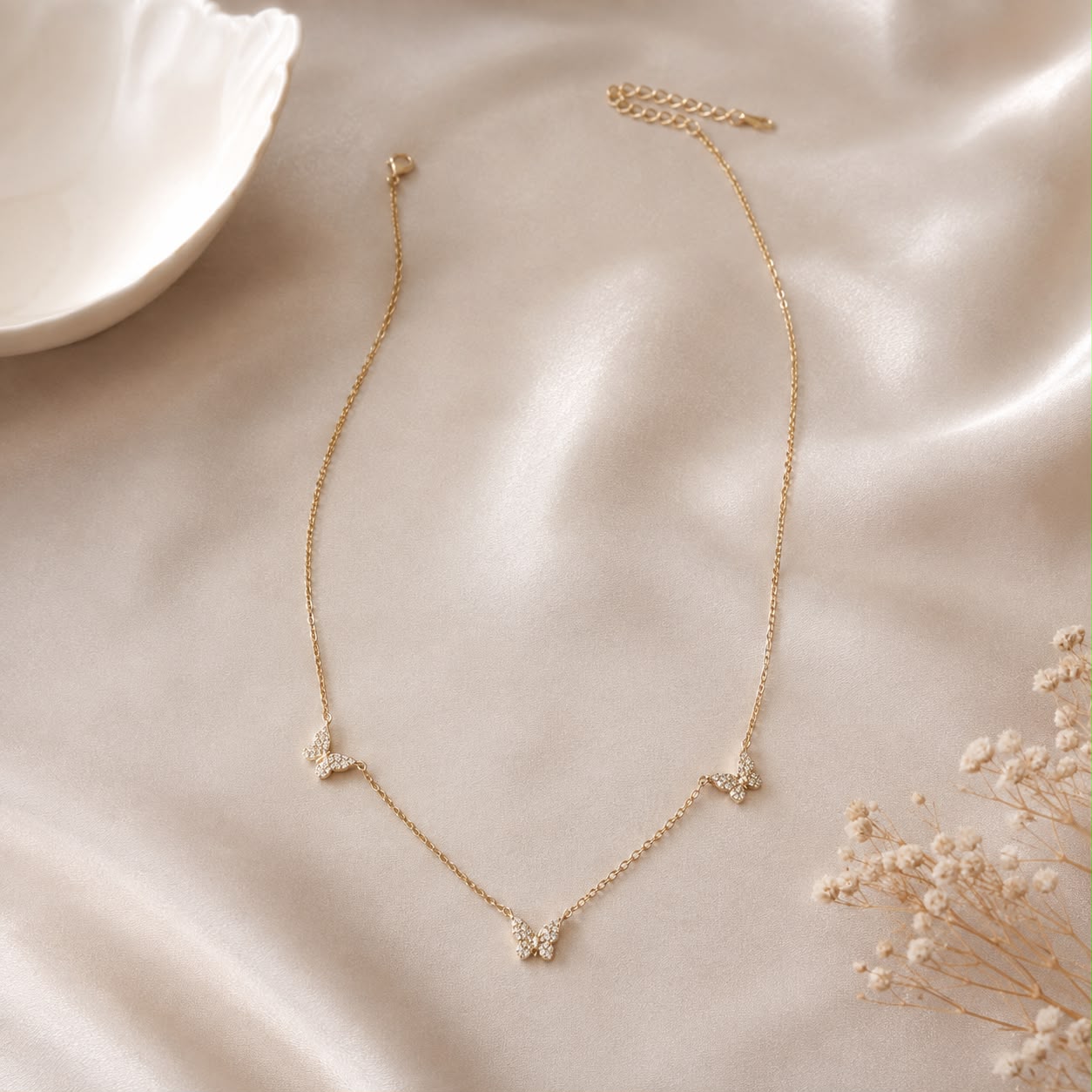 A delicate gold chain necklace with three small butterfly charms, styled on a soft beige satin fabric with a white ceramic dish and dried flowers in the background.