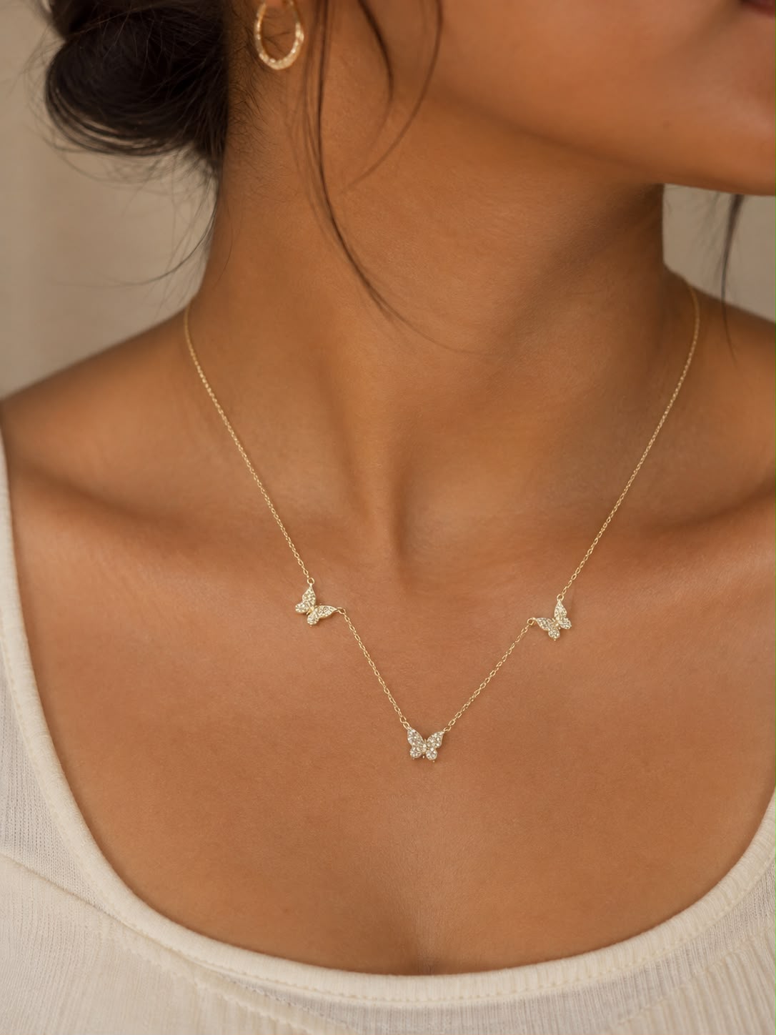 A delicate gold butterfly necklace with three small butterfly charms spaced along a fine chain, worn on a model's neck over a white ribbed top.