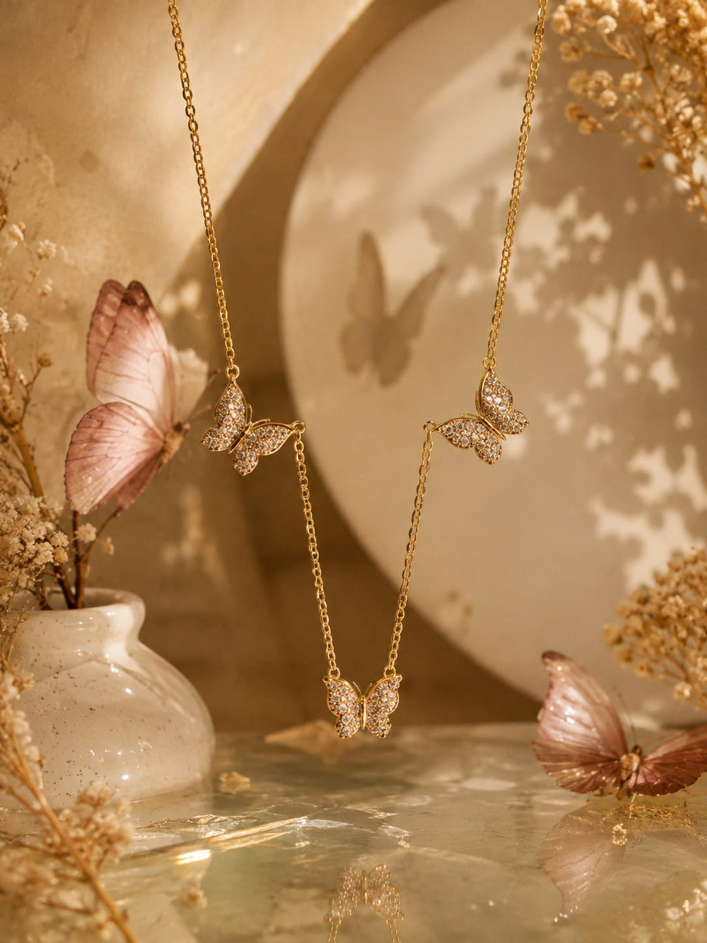 Gold butterfly pendant necklaces displayed on a reflective surface with dried flowers and a soft beige backdrop featuring butterfly shadows.