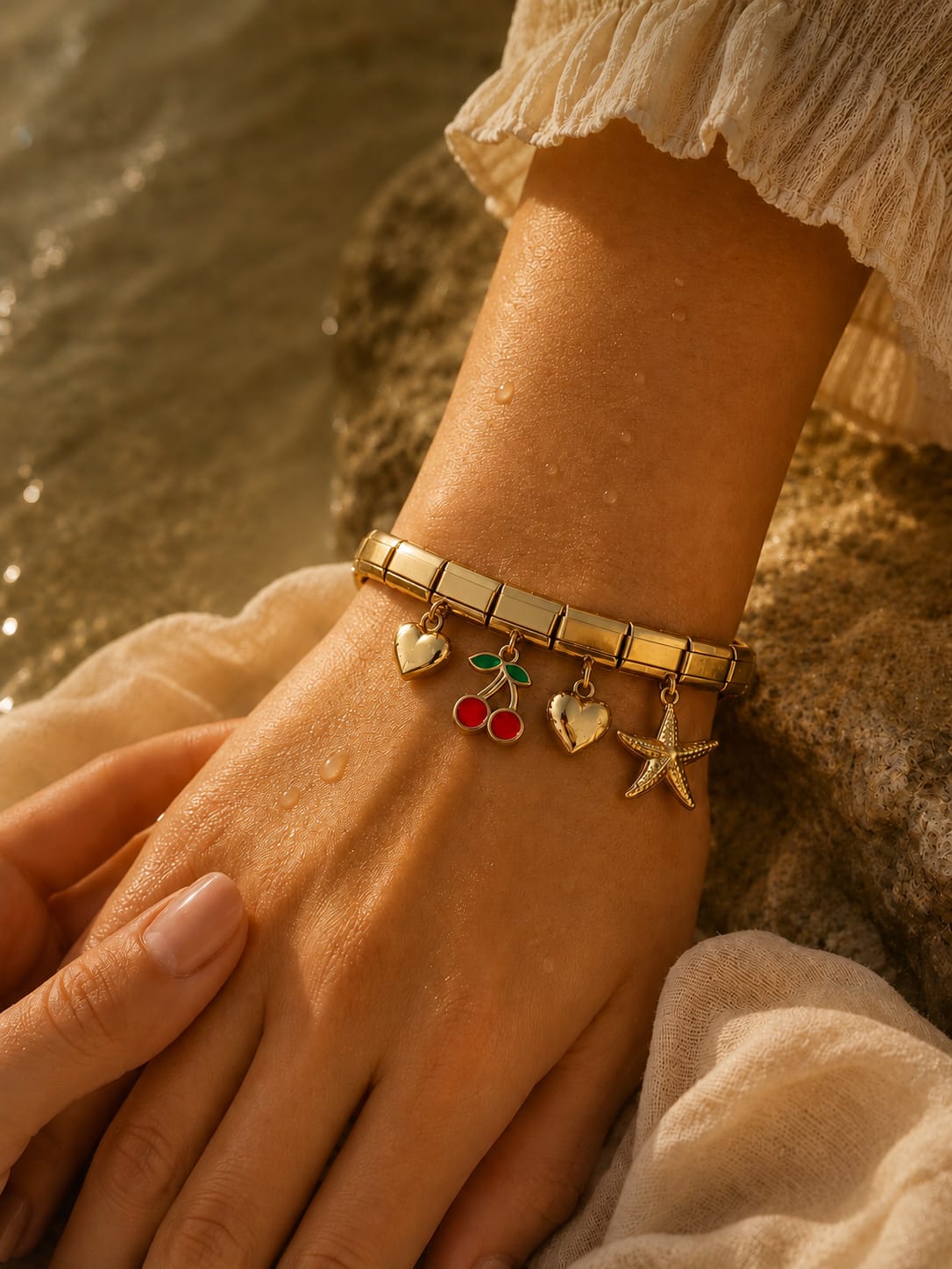 A gold charm bracelet with heart, cherry, and starfish charms worn on a wrist, shown in a warm outdoor setting with water and fabric in the background.