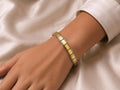 A close-up of a person's wrist wearing a silver-toned tennis bracelet with square yellow-gold inlays, resting on a soft beige satin fabric.