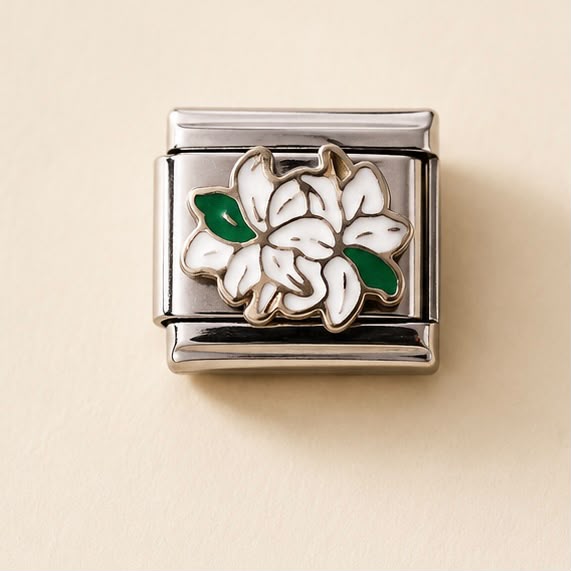 A silver-toned rectangular charm link featuring a white enamel flower with green leaves, displayed on a warm beige background.