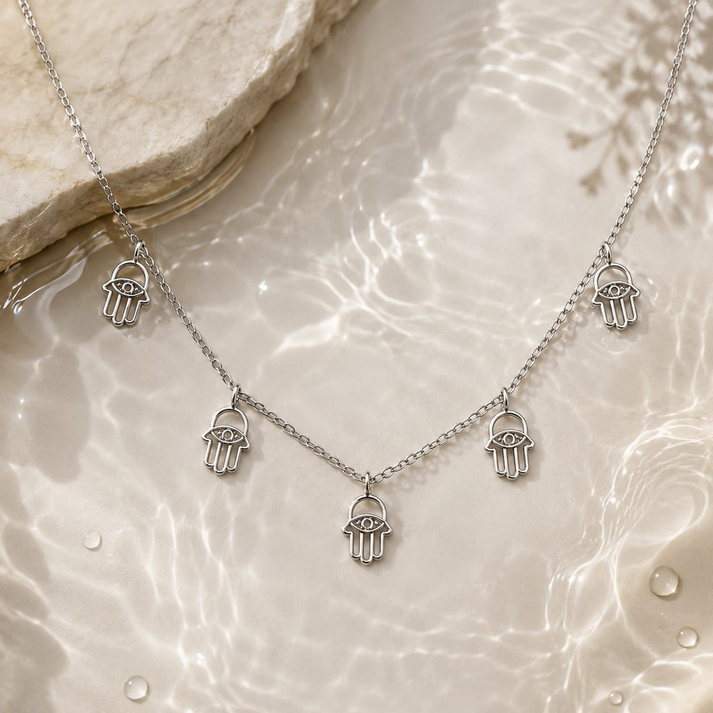 A silver-tone Hamsa hand necklace with five dangling charms, displayed on a glossy, water-textured beige surface with a stone prop in the corner.