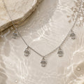 A silver-tone Hamsa hand necklace with five dangling charms, displayed on a glossy, water-textured beige surface with a stone prop in the corner.