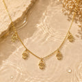 A gold-tone Hamsa hand necklace with an eye charm, styled on a warm beige stone surface with water droplets and dried flowers.