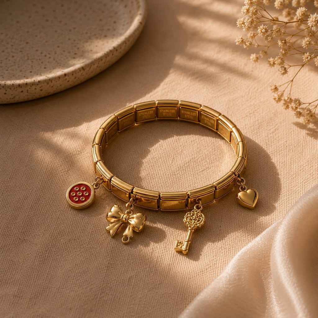 A gold-toned stretch bracelet with three dangling charms, including a red circular charm, a bow charm, and a key charm, displayed on a beige fabric surface with soft shadows.