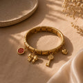 A gold-toned stretch bracelet with three dangling charms, including a red circular charm, a bow charm, and a key charm, displayed on a beige fabric surface with soft shadows.