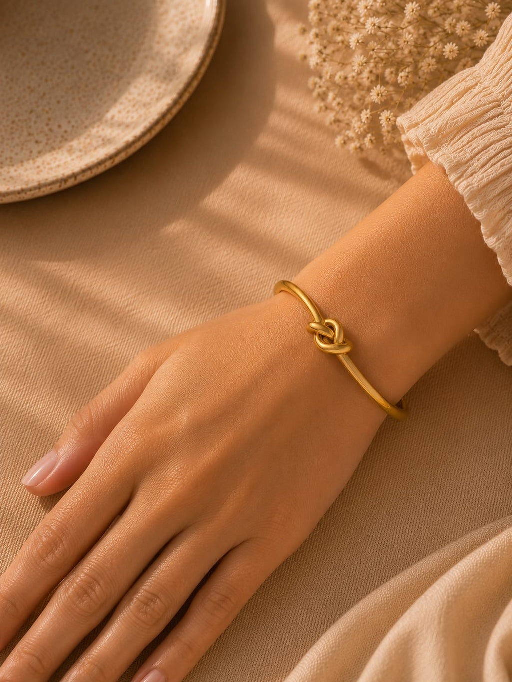 A delicate gold bangle bracelet with a knotted center detail, worn on a woman's wrist. The image is styled with warm, soft lighting, a beige textured fabric background, and a ceramic plate in the upper left corner.
