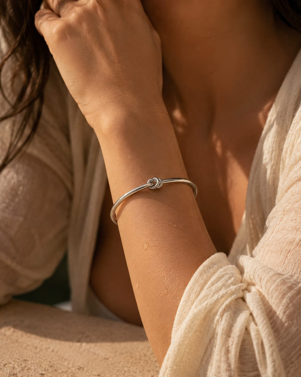 A close-up lifestyle shot of a woman wearing a thin silver bangle bracelet with a small central knot detail, styled with a cream-colored textured top.