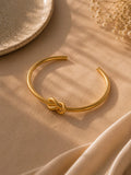 A gold-toned open cuff bracelet with a knotted detail at the front, displayed on a beige fabric surface with soft shadows and dried flowers in the corner.