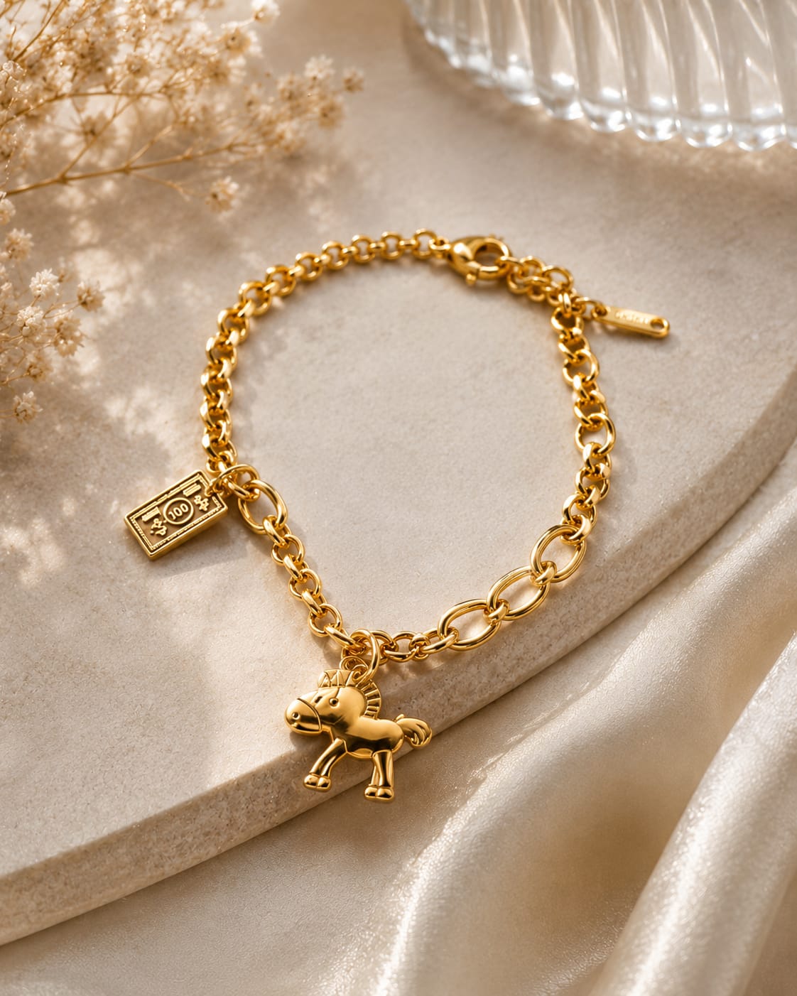 A gold chain bracelet with a small horse charm and a rectangular engraved tag, styled on a beige stone slab with soft satin fabric and dried flowers.