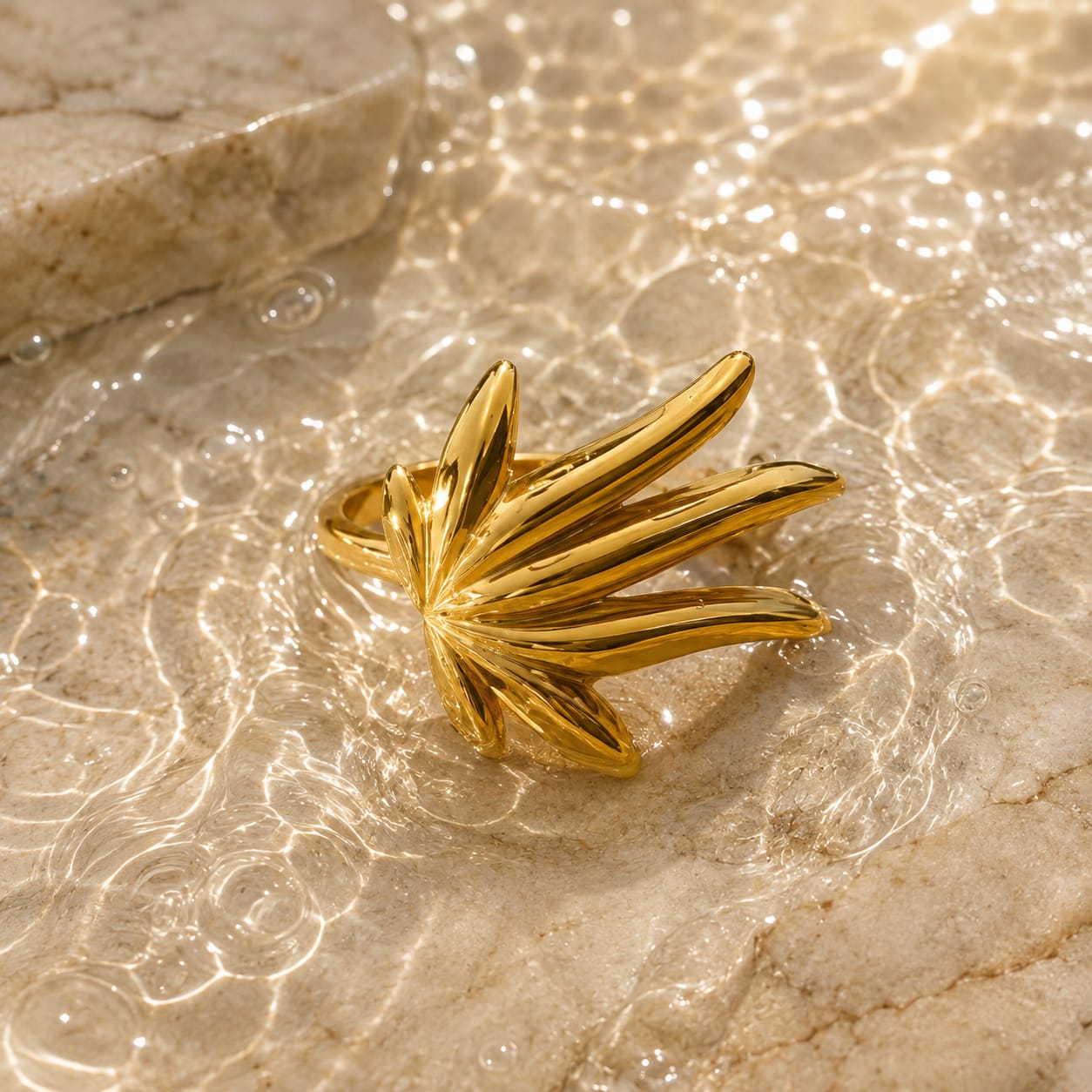A gold ring designed to look like a stylized palm leaf or fan, resting on a textured beige stone surface with shallow water ripples and light reflections.