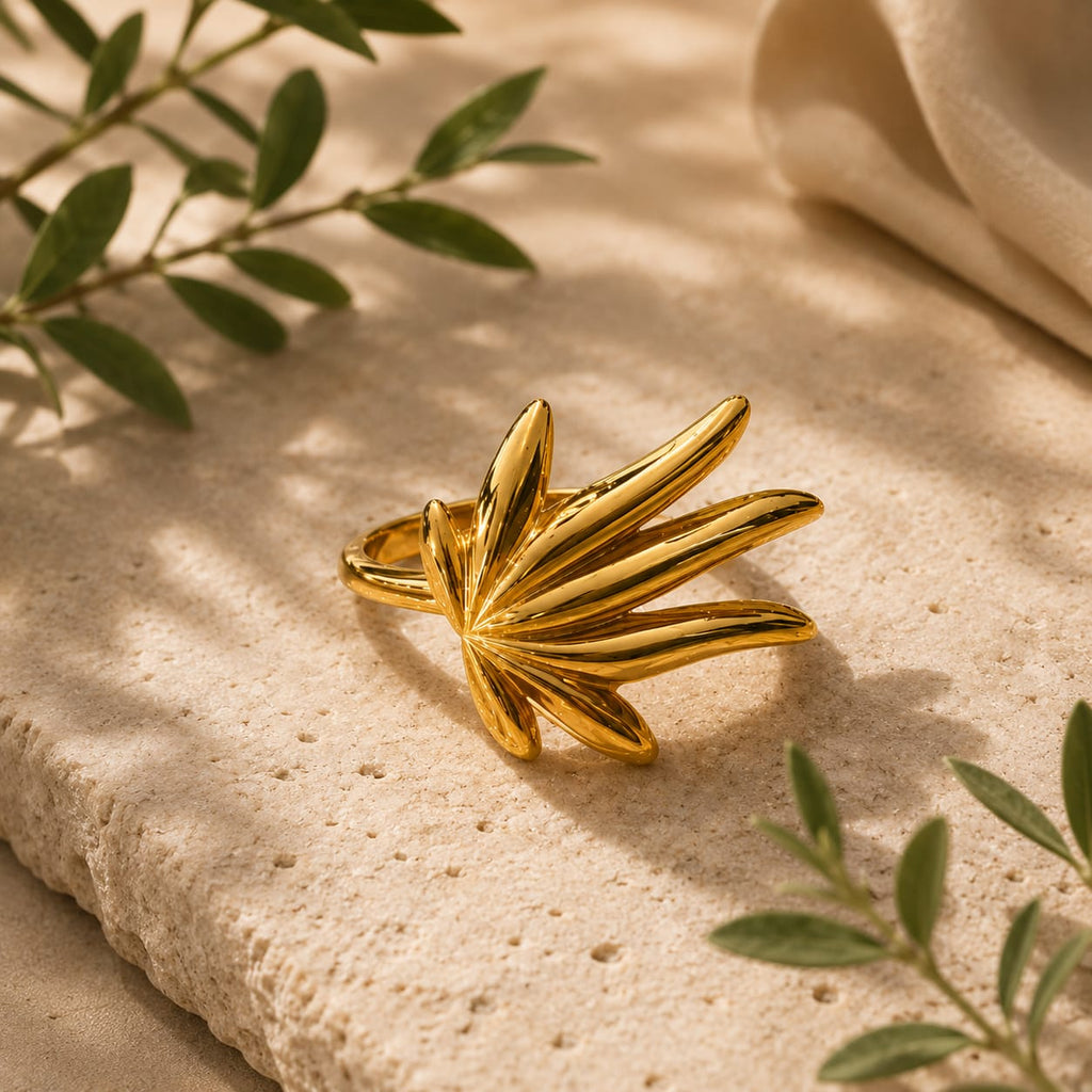 A gold leaf-shaped ring with a polished finish, displayed on a textured beige stone surface with soft shadows and green leaves in the background.