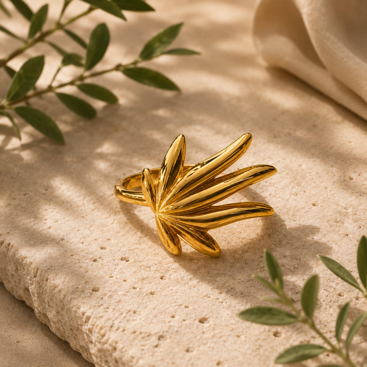 A gold leaf-shaped ring with a polished finish, displayed on a textured beige stone surface with soft shadows and green leaves in the background.