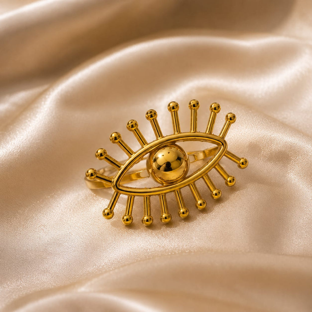 A gold-toned eye-shaped ring with radiating bead-like rays and a polished central sphere, displayed on draped satin fabric.